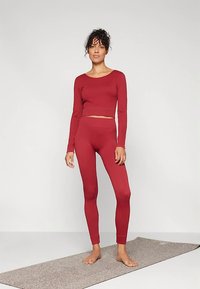 Top vermelho de manga comprida e leggings de cintura alta, design ajustado, textura suave, usado descalço sobre um tapete cinzento contra um fundo simples.
