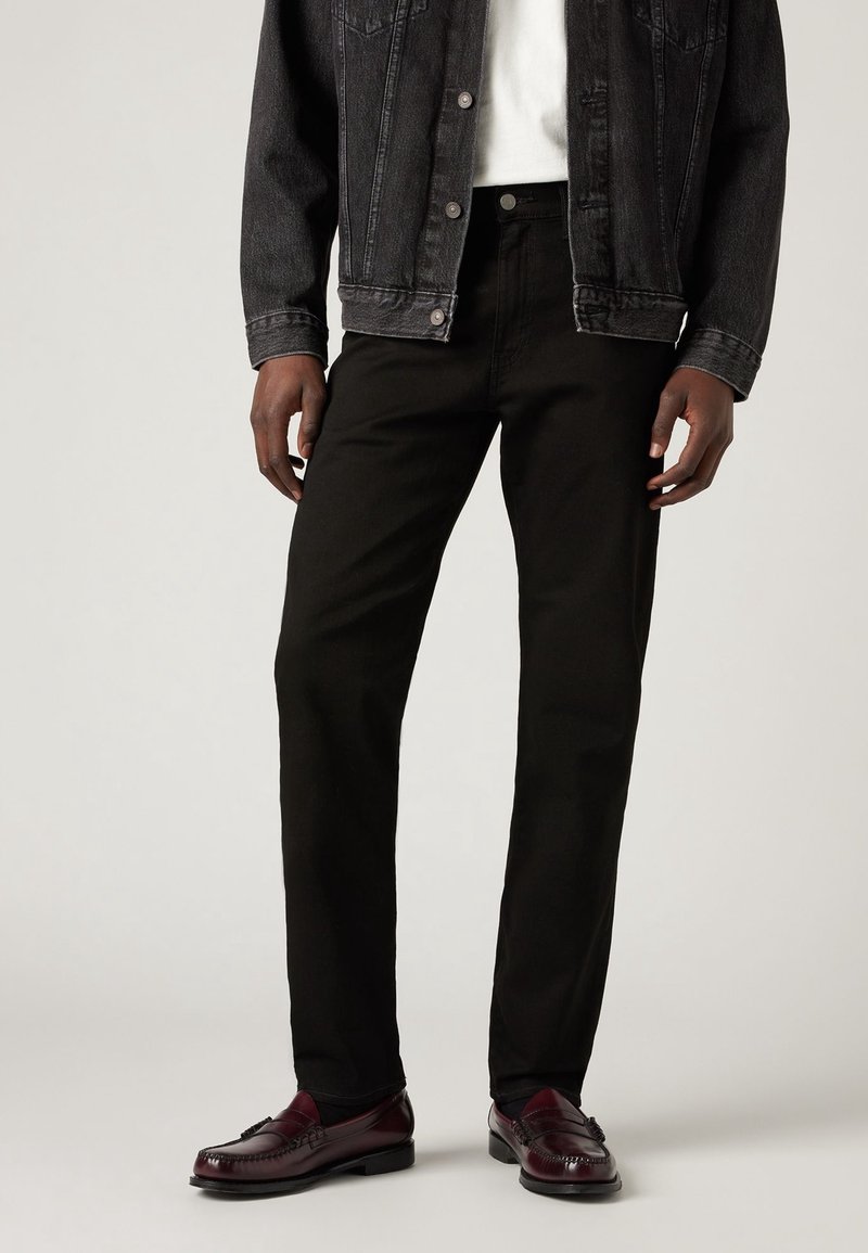 Pantalones vaqueros negros combinados con una chaqueta vaquera negra sobre una camisa blanca. Zapatos de vestir burdeos con detalles de costura completan el conjunto.