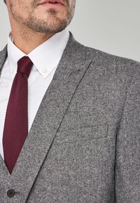 Sacou gri din țesătură tweed cu un finisaj texturat, rever ascuțit și un singur buton. Purtați peste o cămașă albă cu guler și o cravată texturată în nuanță de burgund.