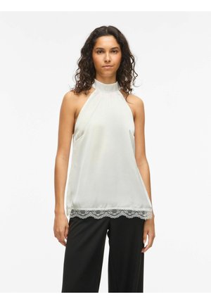 Femme aux cheveux bouclés portant un top blanc sans manches à col halter avec bordure en dentelle, debout devant un fond clair uni, portant un pantalon noir.