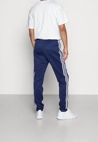 Tmavě modré sportovní kalhoty s bílými bočními pruhy, elastickým pasem a zužujícím se střihem, doplněné o jednoduché bílé tričko a bílé tenisky.