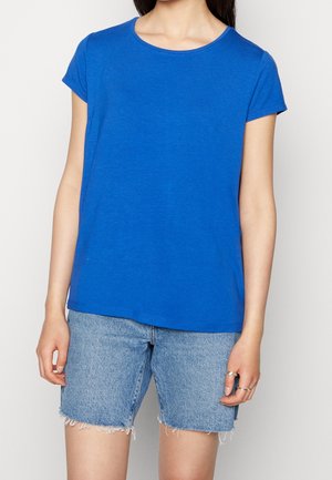 Vrouw draagt een effen blauw T-shirt met korte mouwen en lichtblauwe denim korte broek met rafelige zomen, staand tegen een effen achtergrond.