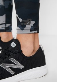 Detailní záběr na kotník oblečený v legínách s maskáčovým vzorem, s logem New Balance, a černou běžeckou obuví New Balance s bílou podrážkou.