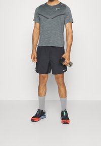 Harmaa Nike lyhythihainen paita, jossa on teksturoitu kuvio, mustat shortsit, harmaat sukat ja mustat lenkkarit, joissa on punaisia ja sinisiä yksityiskohtia, pitämässä vesipulloa.