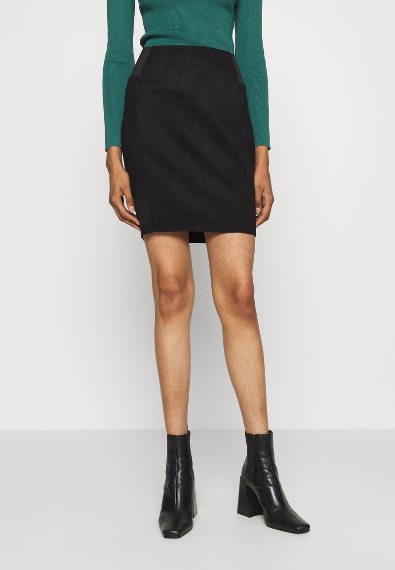 Femme portant une mini-jupe noire, un haut vert à manches longues et des bottines noires à talons, debout devant un fond clair uni.