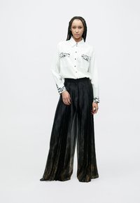 Camicia bianca con dettagli neri a motivo zebra, pantaloni lunghi neri plissettati con una leggera lucentezza, vestibilità sartoriale con bottoni visibili.