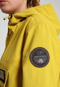Gele windjack met capuchon, met een zwarte ronde patch op de mouw en een groot zak aan de voorkant.