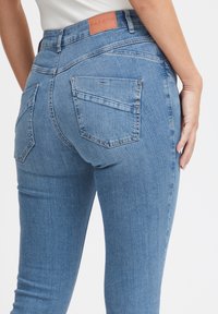 Mršavi traperice svijetloplave boje, s kožnom oznakom, dva stražnja džepa i metalnim ukrasnim gumbima na desnom džepu.