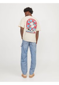 Beige bomulds T-shirt med et farverigt grafik af en kaffekande og en kop på ryggen. Sammen med lyseblå jeans og brune sko.