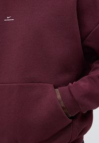 Σκούρα κόκκινο φούτερ Nike με μικρό λευκό λογότυπο και κείμενο "NIKE STANDARD ISSUE" στην αριστερή πλευρά του στήθους, χέρι μερικώς στην μπροστινή τσέπη.