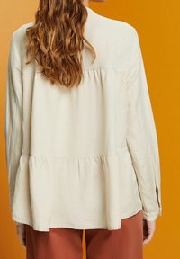 Blusa de manga larga color crema con un dobladillo acampanado, hecha de tejido suave. Presenta aberturas laterales y un escote redondo sobre un fondo naranja.