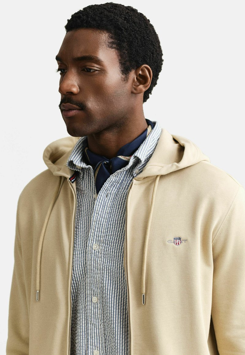 Junger Mann mit kurzen lockigen Haaren und Schnurrbart, der einen beigen Hoodie über einem blau gestreiften Hemd mit Knöpfen und einem dunklen Halstuch trägt, nach links blickend.