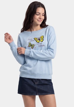 Ung kvinde iført en lyseblå sweatshirt med farverige sommerfugleprint og tekst, kombineret med et mørkt denimskørt.