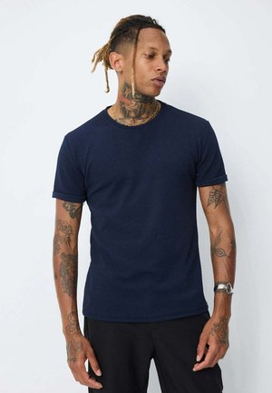 T-shirt bleu marine à manches courtes avec un tissu texturé, col rond et revers retroussés. Porté avec un pantalon noir, présente des tatouages visibles.
