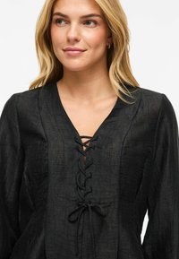 Svart bluse med lange ermer og teksturert finish, med V-hals og snøring foran. Mykt stoff med avslappet passform.