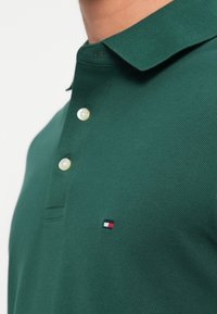 Groene polo met twee witte knopen en een klein geborduurd logo in rood, wit en blauw op de borst.