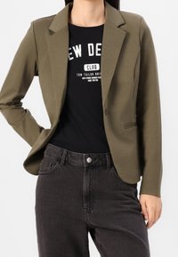 Olijfgroen blazer met een op maat gemaakte stijl, sluiting met één knop en notch revers, gedragen over een zwart T-shirt met een grafisch ontwerp en donkere jeans.