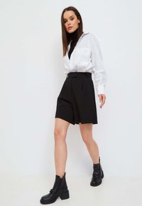 Weißes Hemd mit Knöpfen und zwei Brusttaschen, kombiniert mit hochgeschnittenen schwarzen Plissee-Shorts und schwarzen Ankle Boots mit klobigen Sohlen.