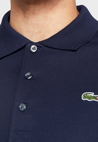 Detailní záběr na osobu v tmavě modrém polo tričku se třemi knoflíky a zeleným krokodýlím logem na hrudi.