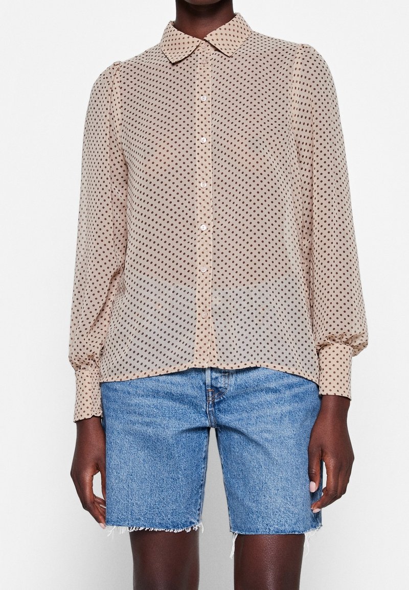 Blusa beige a pois con colletto, apertura frontale con bottoni e maniche lunghe a sbuffo, abbinata a pantaloni corti di jeans sfrangiati.