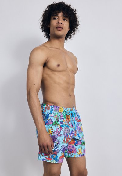 Oben ohne man mit lockigem Haar in bunten Badehosen mit einem Muster von Unterwasserlebewesen, steht vor einem schlichten Hintergrund.
