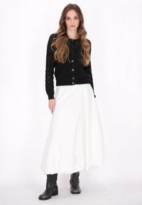 Cardigan nero a maglia con un motivo in pizzo, con bottoni sulla parte anteriore; abbinato a una gonna bianca fluida e stivaletti neri alla caviglia.