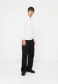 White long-sleeve pullover with a zip collar, paired with black cargo pants featuring side pockets and a relaxed fit. Black slip-on shoes.