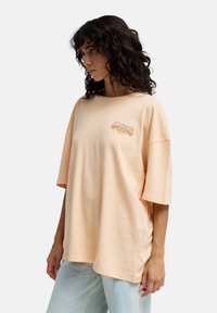 Ροδακινί υπερμεγέθη μπλουζάκι από βαμβάκι, με κοντά μανίκια και λογότυπο με floral κεντήματα στο στήθος.