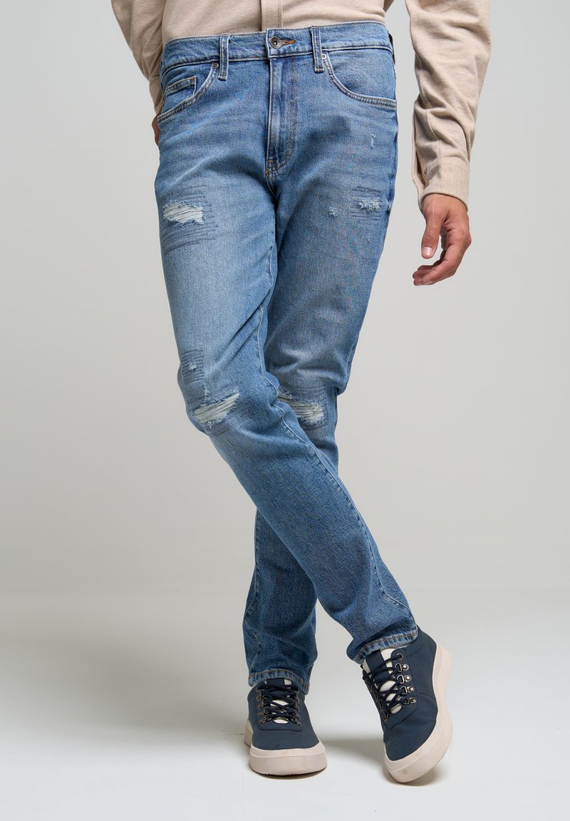 Heled sinine teksapüksid, millel on kulumise üksikasjad, sirge lõige, standardne viie tasku disain, kandmas tumesiniseid tennisetossud, millel on hallid aktsendid.