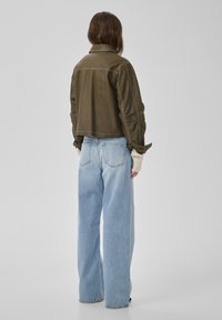 Femeie văzută din spate, purtând o geacă de piele verde măsliniu, blugi evazați bleu deschis și mănuși fără degete de culoare crem.