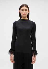 Žena oblečená v priliehavom čiernom rebrovanom tričku s dlhým rukávom a perovými manžetami a čiernych širokých nohaviciach, stojaca pred jednoduchým bielym pozadím.