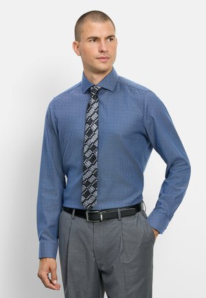 Man draagt een blauw patroon lang mouw shirt met een zwart-witte geometrische patroon stropdas en grijze broek, staat met één hand in de zak.
