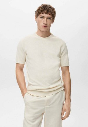 Beige, short-sleeve knit sweater with a ribbed neckline and textured fabric. Worn with light beige trousers, showcasing a minimalist design.