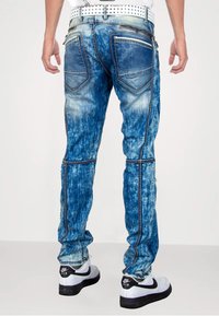 Blauwe denim jeans met een vervaagd ontwerp, contrasterende stiksels, twee achterzakken en een ritsdetail aan de zijkanten. Zwart-witte sneakers gedragen.