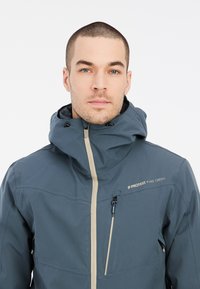 Blauwe skijas met een hoge kraag, rits aan de voorkant en een trekkoord kap. Bevat een voorzak met contrasterende rits en logodetail.