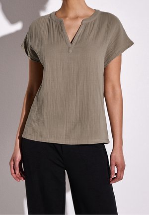 Vrouw draagt een taupe korte mouwen textuurt blouse met een ingesneden halslijn en zwarte broek, staand tegen een lichte achtergrond.