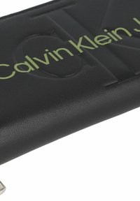 Portafoglio in pelle nera con logo "Calvin Klein" in verde a rilievo. Presenta una superficie testurizzata e una forma rettangolare semplice. Finiture in metallo argentato.