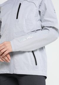 Jasnoszara kurtka w kratę z czarnymi suwakami, ręce skrzyżowane, logo "THE NORTH FACE" na rękawie i ramieniu.