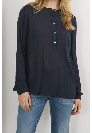 Femme portant un chemisier ample bleu marine à boutons avec manches longues et un jean en denim bleu clair, debout devant un fond uni.