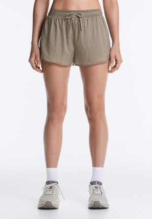 Frau trägt beige Shorts mit Kordelzug, weiße Knöchelsocken und hellgraue Laufschuhe, steht mit entspannten Armen neben dem Körper.