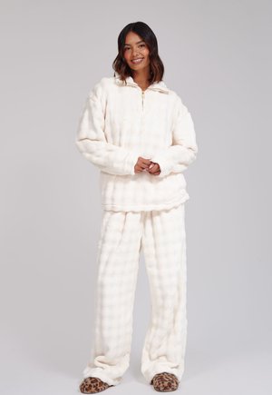Femme debout et souriante, portant un pull polaire blanc doux et un pantalon avec des chaussons à motif léopard sur un fond uni.