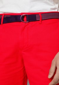Gros plan sur une personne portant un pantalon rouge vif avec une ceinture rayée noire, rouge et blanche, fixée par une boucle métallique en forme de D.