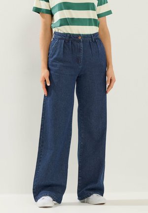 Person iført blå jeans med høj talje og vide ben med folder, en grøn- og hvidstribet kortærmet skjorte og hvide sneakers, står.