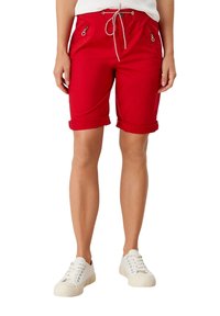 Pantalones cortos rojos con cinturilla ajustable mediante cordón, puños doblados y bolsillos con cremallera. Fabricados con un tejido resistente y de textura suave. Combinados con zapatillas blancas.