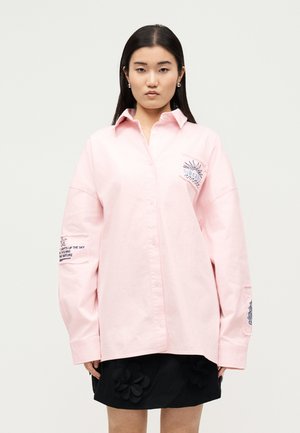 Γυναίκα που φοράει υπερμεγέθη ανοιχτό ροζ πουκάμισο με κεντημένο ήλιο και κείμενο, σε συνδυασμό με μαύρη φούστα με κεντήματα λουλουδιών.