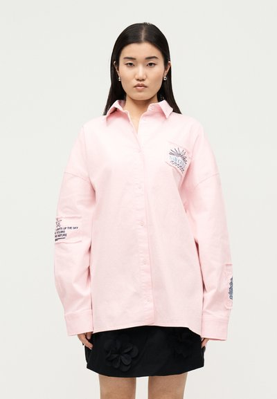 Femme portant une chemise rose clair oversize avec un soleil brodé et du texte, associée à une jupe noire ornée d'appliqués floraux.