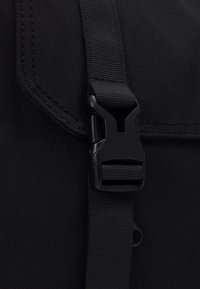 Sangle en nylon noir avec une boucle en plastique noire, présentant un tissu texturé et des bords cousus, fixée à un grand sac noir.