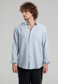 Giovane uomo con capelli ricci, che indossa una camicia a righe azzurro chiaro, con maniche lunghe e bottoni, e pantaloni neri, che guarda a sinistra.
