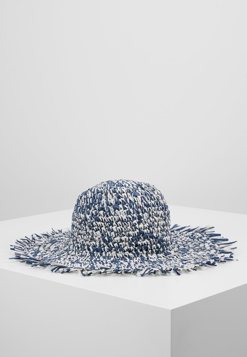 Chapeau tissé bleu et blanc à large bord avec des bords effilochés, exposé sur un piédestal blanc sur un fond uni.