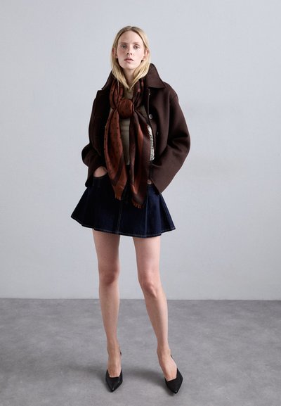 Manteau en laine marron par-dessus un haut beige, associé à une jupe en denim foncé. Une écharpe à motifs marron drape le cou. Escarpins noirs à bout pointu.